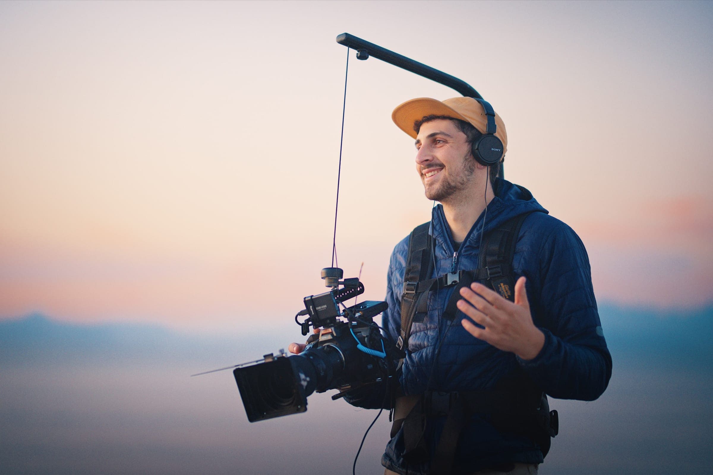 A camera operator in a cap and headphones holds a cinema camera on a support rig, against a pastel sunset sky.